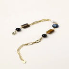 Gold chain bracelet with tiger's eye, obsidian, pearl, and iridescent abalone stones on cream background.