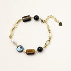 Gold chain bracelet with tiger's eye, obsidian, pearl, and iridescent abalone stones on cream background.