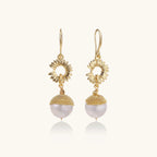 Gold sunburst charm earrings with baroque pearl drops on French hooks against cream background