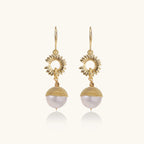 Gold sunburst charm earrings with baroque pearl drops on French hooks against cream background