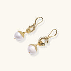 Gold sunburst charm earrings with baroque pearl drops on French hooks against cream background