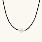Delicate necklace with black blue sandstone beads and baroque pearl center on white background with gold accents.
