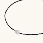 Delicate necklace with black blue sandstone beads and baroque pearl center on white background with gold accents.