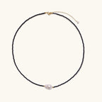 Delicate necklace with black blue sandstone beads and baroque pearl center on white background with gold accents.