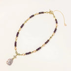 Gold necklace with iridescent baroque pearl pendant, purple amethyst beads, and crystal star charm on cream background
