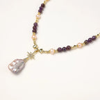 Gold necklace with iridescent baroque pearl pendant, purple amethyst beads, and crystal star charm on cream background