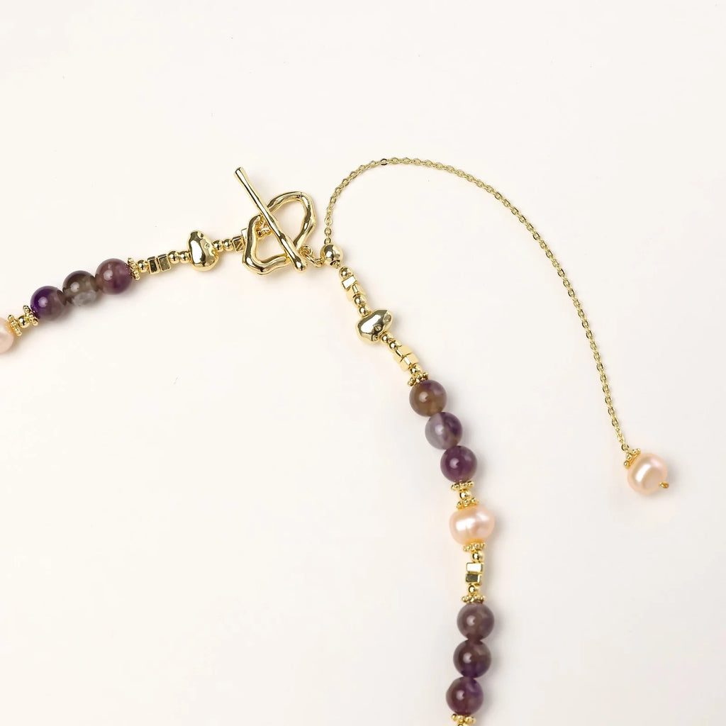 Gold necklace with iridescent baroque pearl pendant, purple amethyst beads, and crystal star charm on cream background