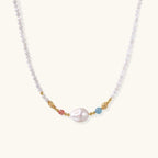 Delicate white beaded necklace with baroque pearl center, gold accents, and coral and aqua gemstones on cream background.