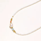 Delicate white beaded necklace with baroque pearl center, gold accents, and coral and aqua gemstones on cream background.