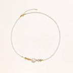 Delicate white beaded necklace with baroque pearl center, gold accents, and coral and aqua gemstones on cream background.