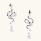 Silver snake-shaped drop earrings with crystal accents and teardrop stone dangles on cream background.