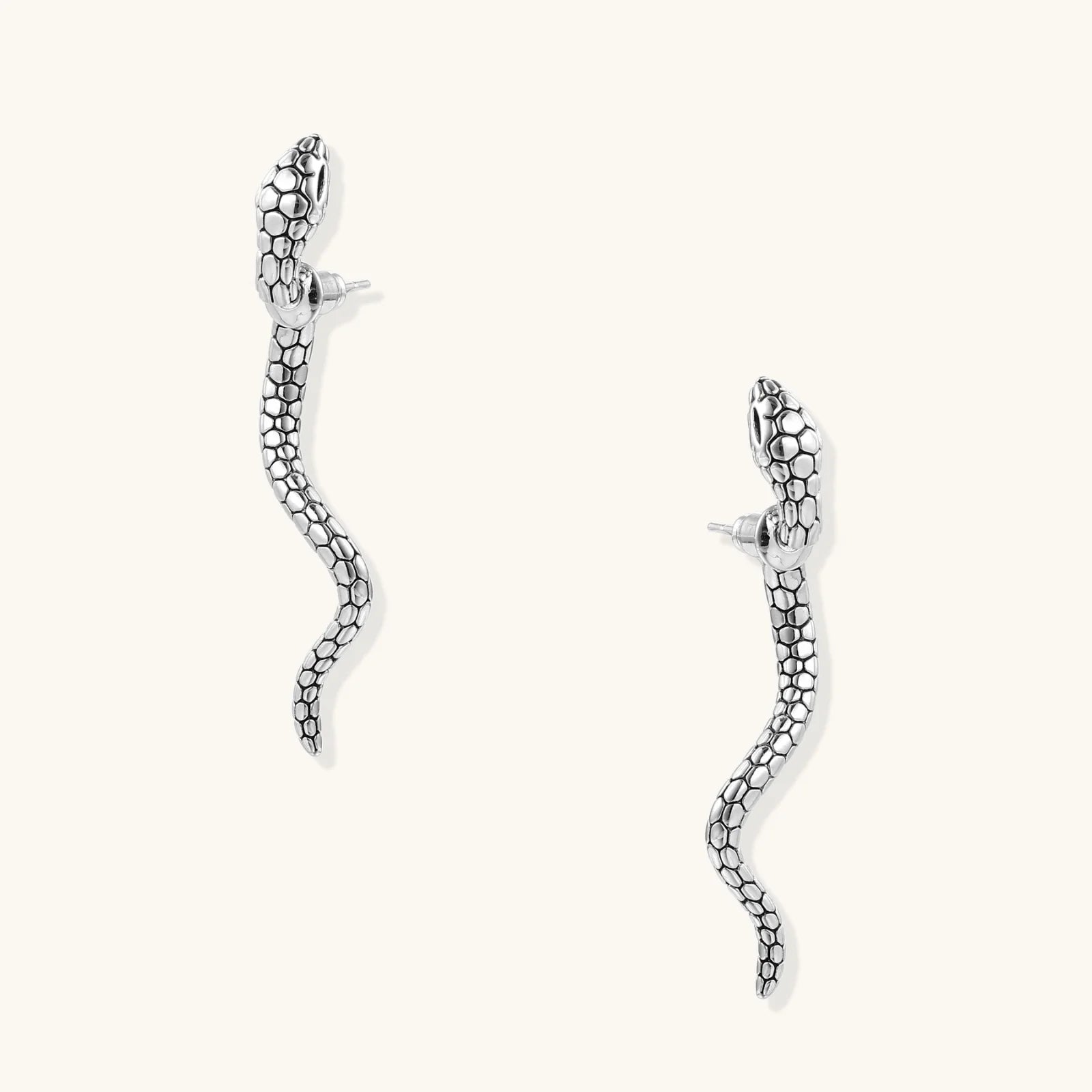 Silver-toned serpent-shaped dangle earrings with detailed scale texture curving in S-shape on beige background