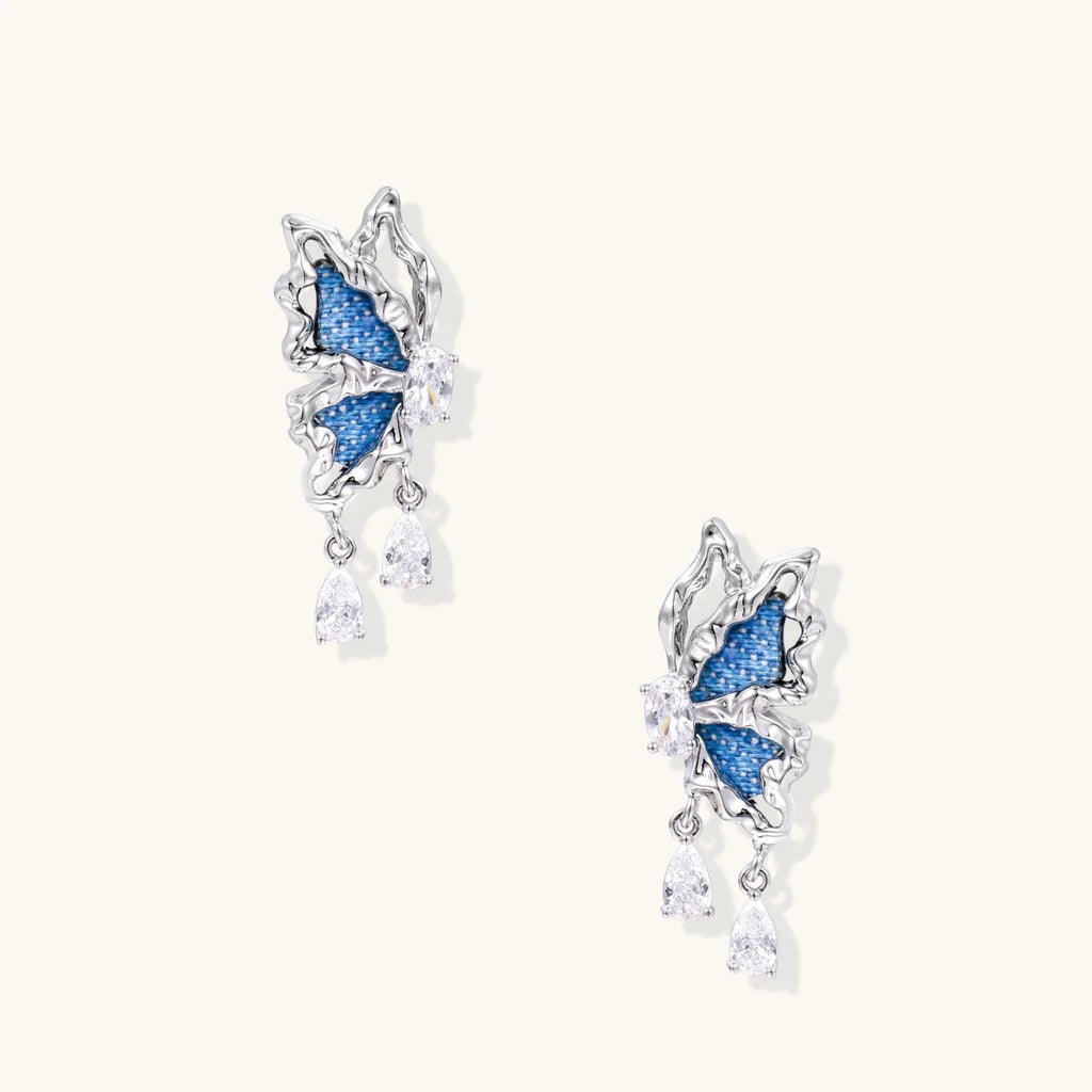 Silver butterfly earrings with blue denim fabric wings and dangling teardrop crystals on cream background