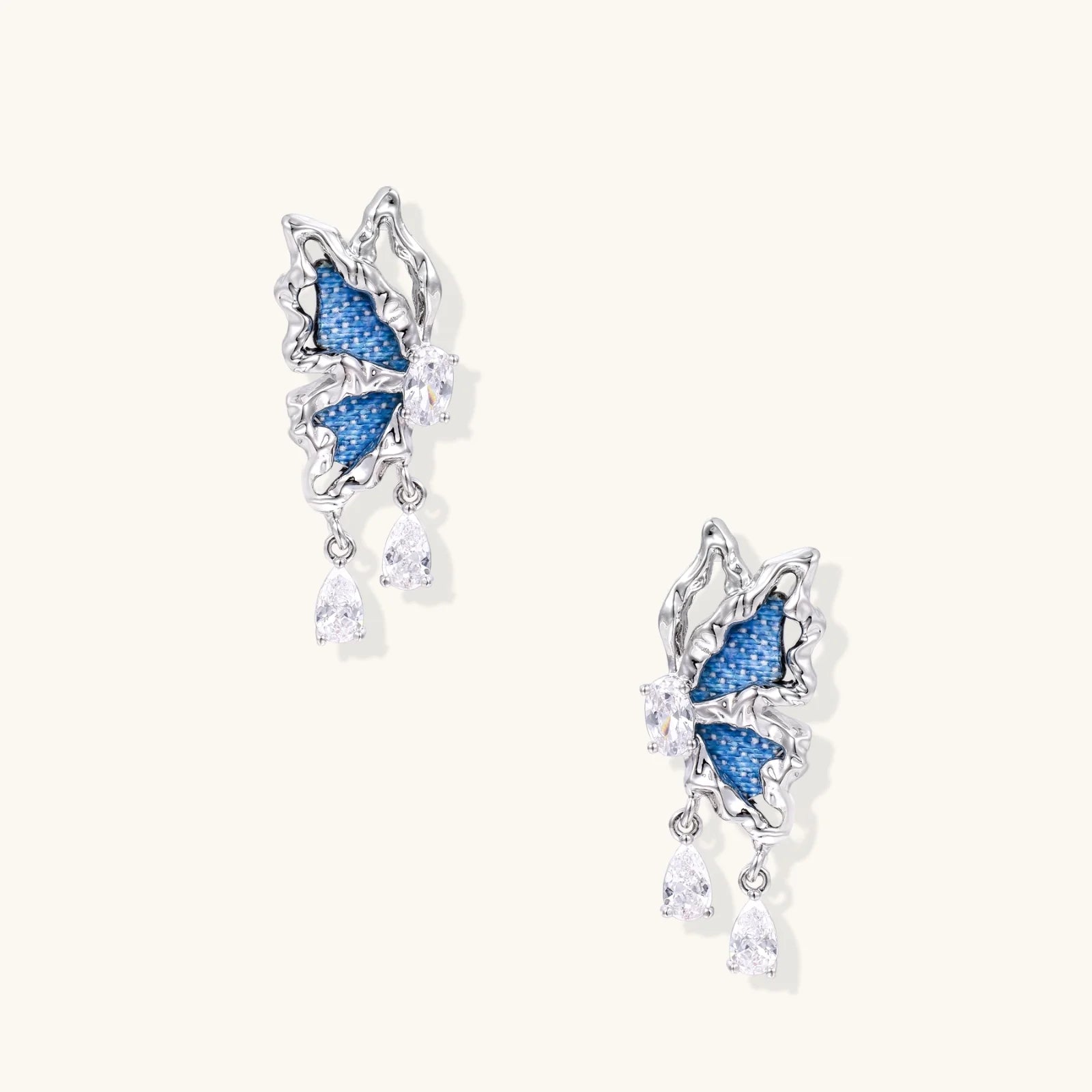 Silver butterfly earrings with blue denim fabric wings and dangling teardrop crystals on cream background
