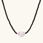 Beaded black agate necklace with iridescent baroque freshwater pearl centerpiece on cream background.