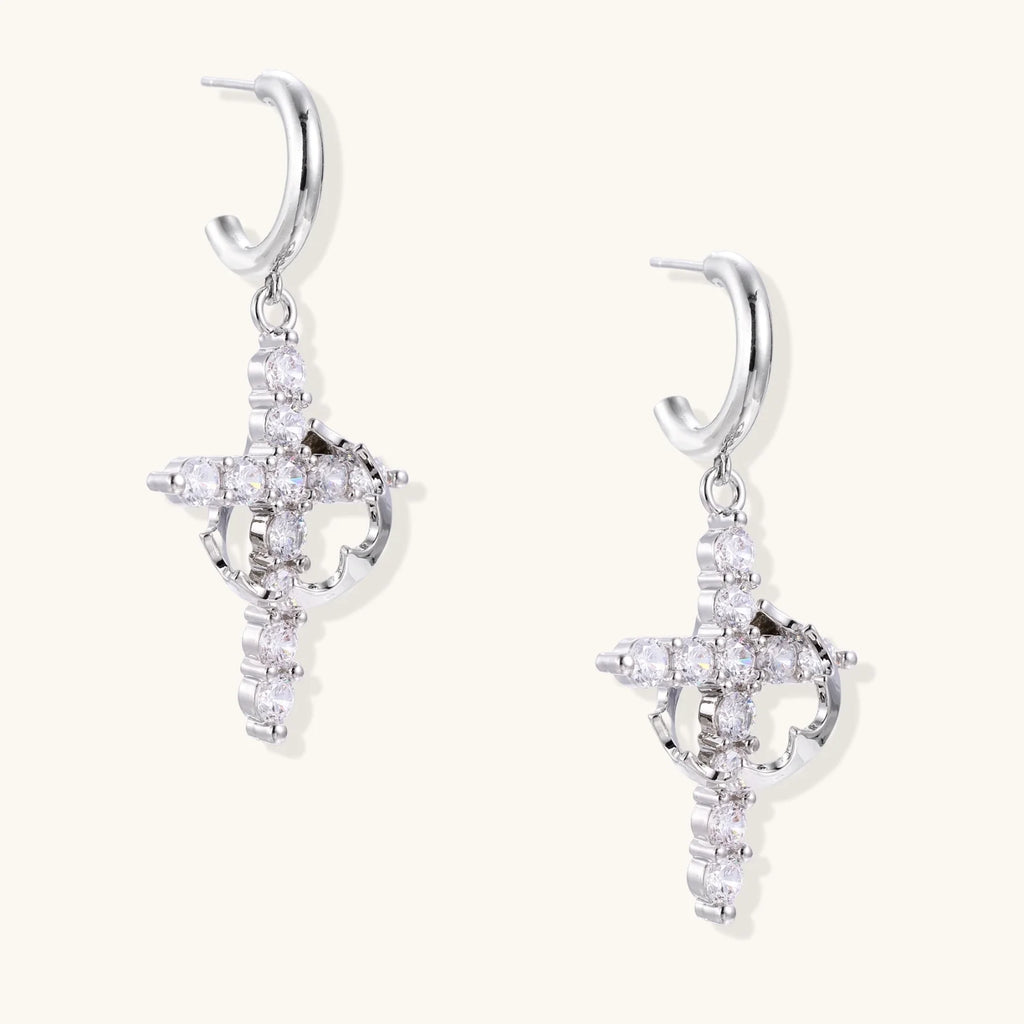White gold-plated hoop earrings with sparkling zirconia cross charms dangling against cream background