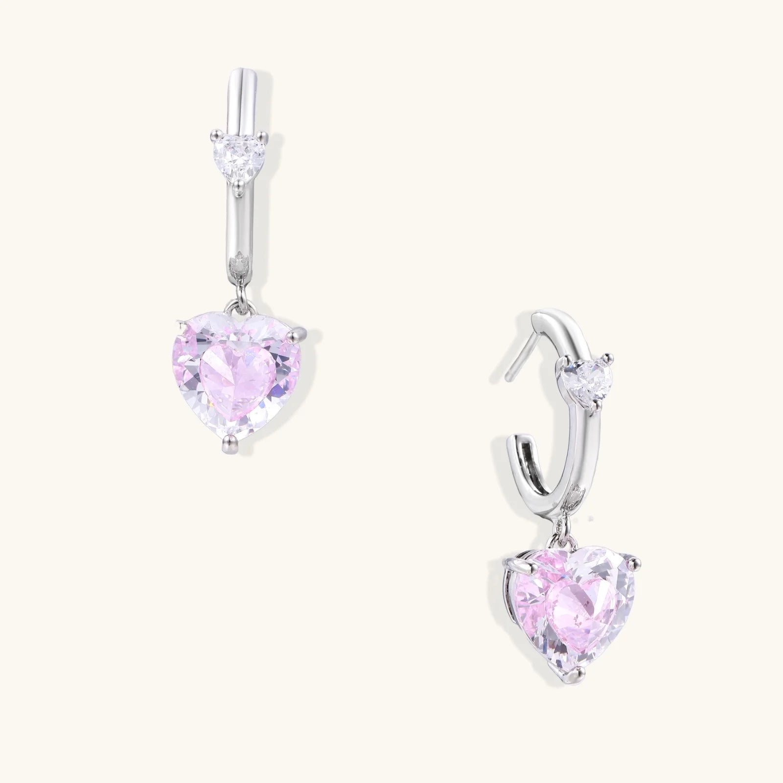 Silver huggie hoops with pink heart-cut zirconia pendants on white background, eco-friendly white gold plated brass