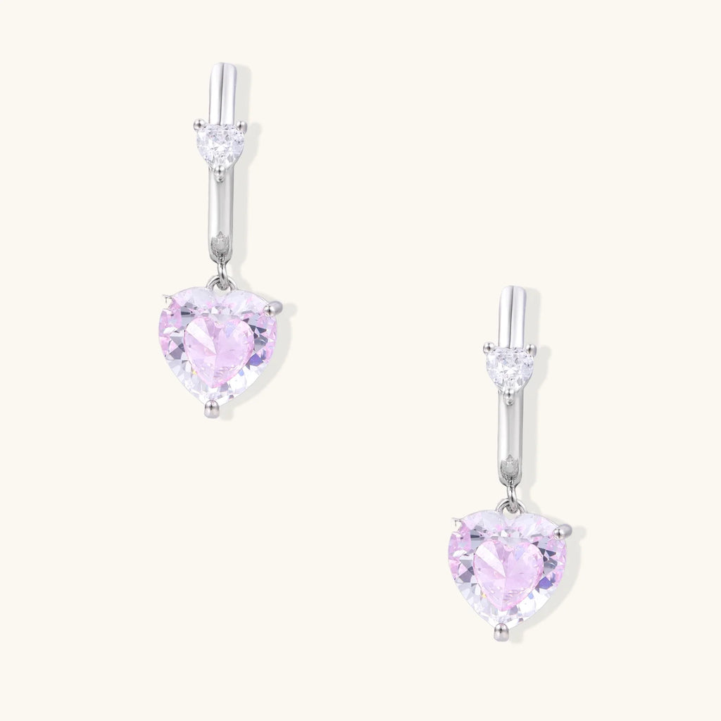 Silver huggie hoops with pink heart-cut zirconia pendants on white background, eco-friendly white gold plated brass