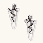 Silver dangle earrings with sculptural serpent coiled around cross shape and delicate chain accents on cream background