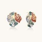 Artistic monstera-shaped earrings with teal, coral, and cream enamel brushstrokes, gold trim, and white pearl accent
