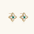 Gold quatrefoil stud earrings with white mother-of-pearl petals and emerald green gemstone center on beige background.