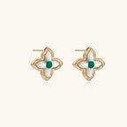 Gold quatrefoil stud earrings with white mother-of-pearl petals and emerald green gemstone center on beige background.
