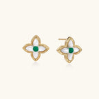 Gold quatrefoil stud earrings with white mother-of-pearl petals and emerald green gemstone center on beige background.