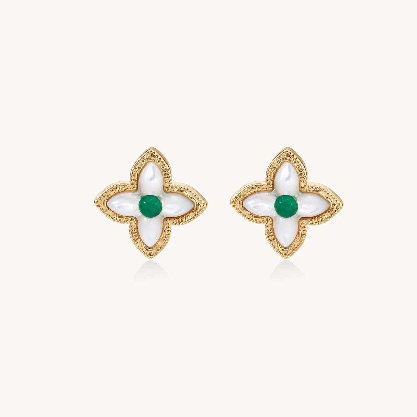 Gold quatrefoil stud earrings with white mother-of-pearl petals and emerald green gemstone center on beige background.