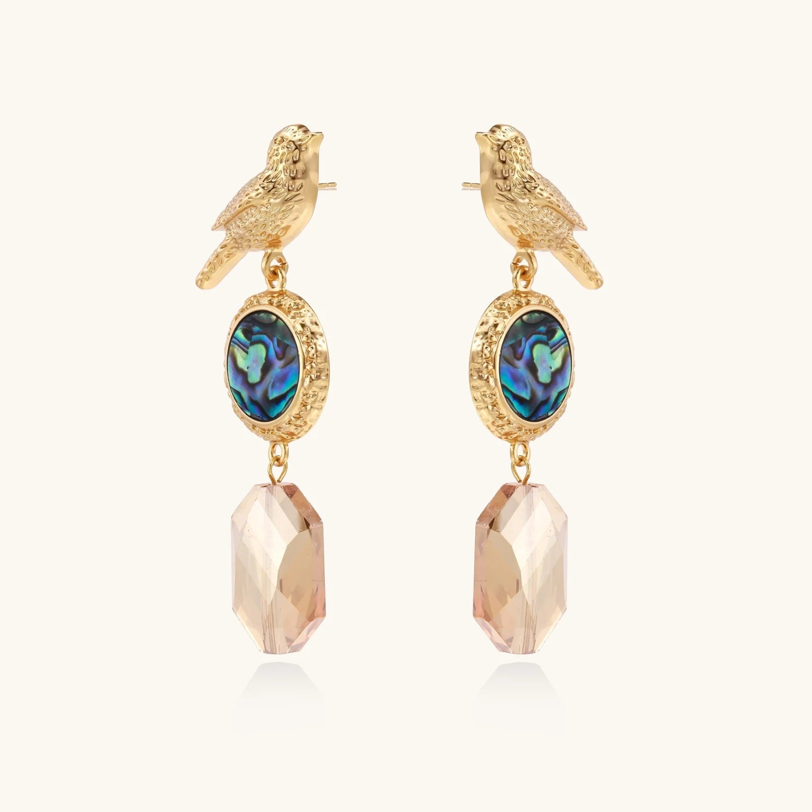 Gold-plated bird stud earrings with iridescent abalone shell medallions and champagne crystal drops on cream background