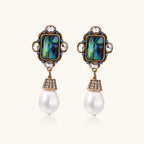 Ornate gold-plated earrings with iridescent abalone shell, crystals, blue stones, and white pearl drops on cream background.