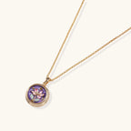 Gold-plated circular pendant with hand-painted purple enamel floral design on delicate chain against cream background