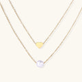 Two delicate gold-plated chains with heart charm and white pearl bead on cream background, minimal layered design.