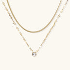 Layered gold necklace with herringbone and link chains, small bezel-set crystal pendant on cream background