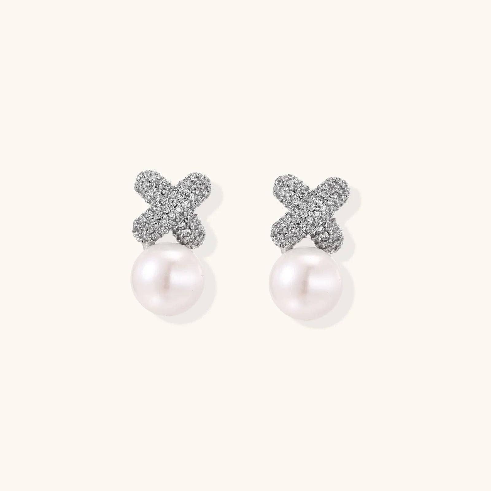 White gold plated studs featuring pavé X-shaped tops with freshwater pearl drops on cream background.