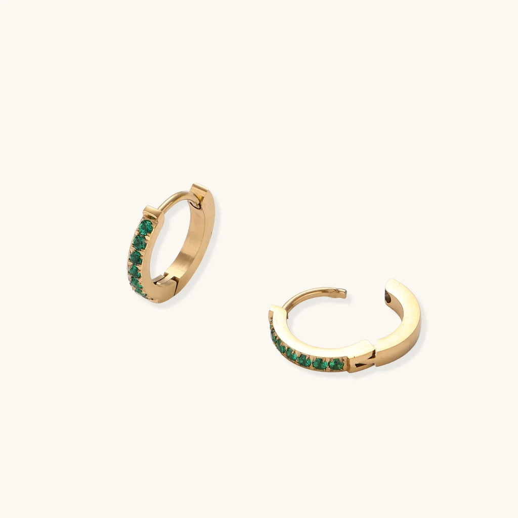 Pair of small gold huggie hoop earrings with bright green cubic zirconia stones on cream background