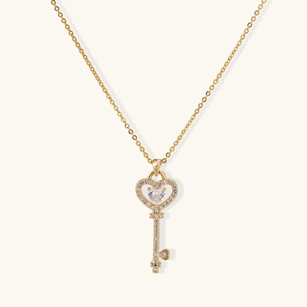 Delicate gold key pendant with heart-shaped bow and crystal pavé detailing on fine chain against cream background.