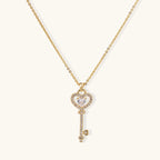 Delicate gold key pendant with heart-shaped bow and crystal pavé detailing on fine chain against cream background.