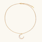 Gold crescent moon pendant with graduated white pearls on delicate chain against cream background