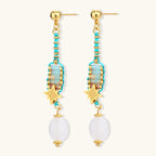 Gold-plated dangle earrings with turquoise threading, star charms, and white pearl beads against cream background.