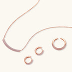 Rose gold jewelry set with pavé cubic zirconia stones including curved necklace, hoop earrings, and adjustable ring on cream background.