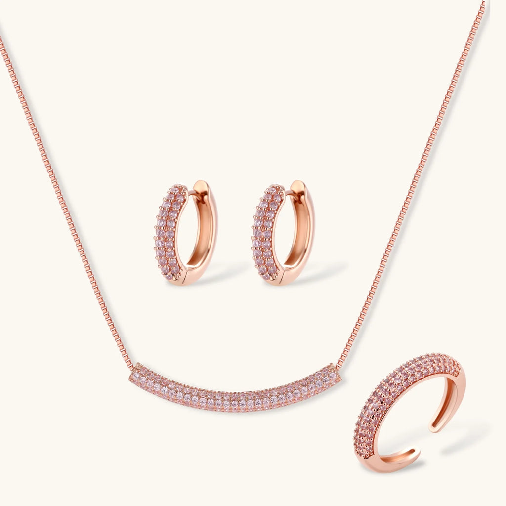 Rose gold jewelry set with pavé cubic zirconia stones including curved necklace, hoop earrings, and adjustable ring on cream background.
